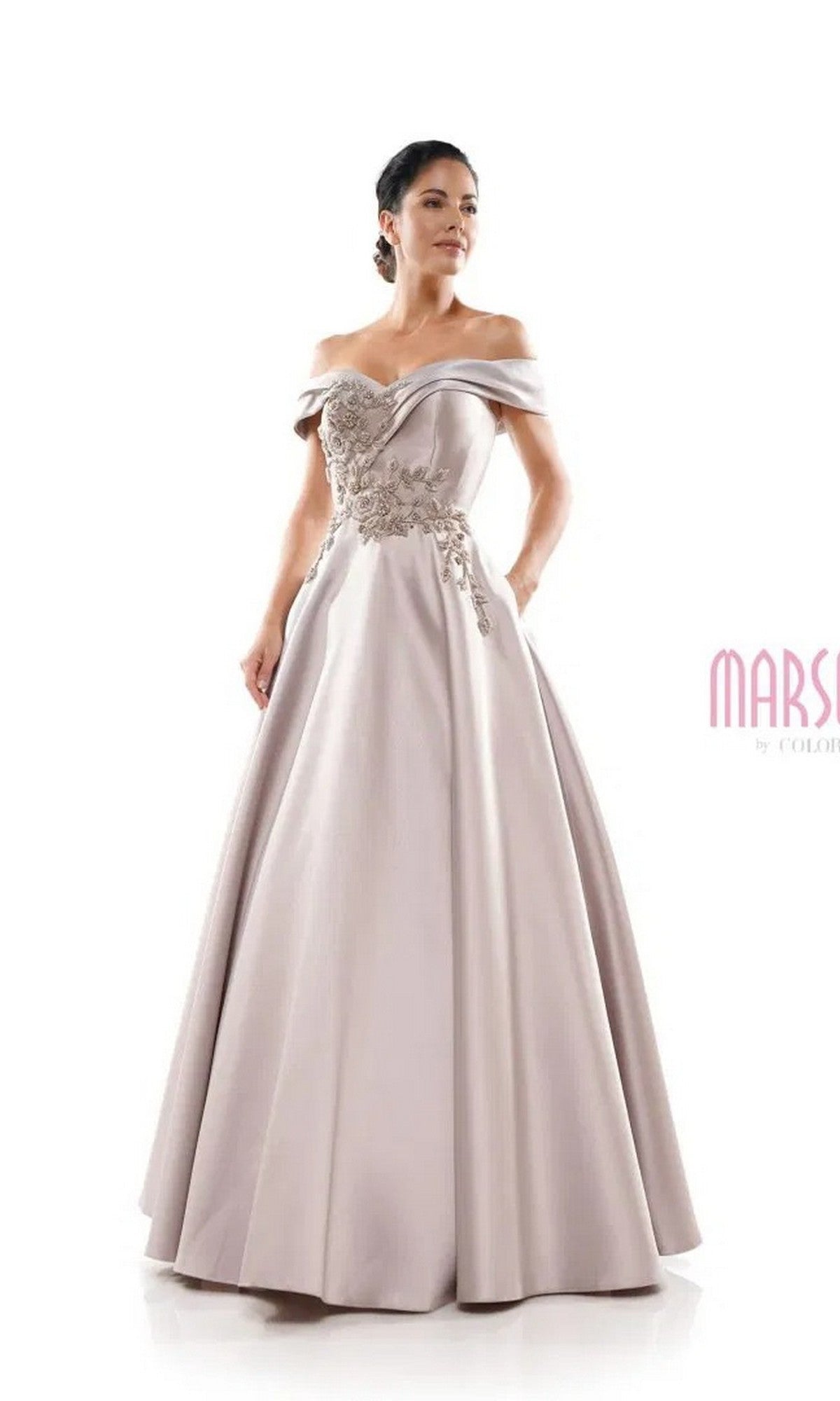 A woman wears the Marsoni MV1008 long formal dress in taupe, featuring an off-the-shoulder design, floor-length skirt, floral embroidery, and delicate beaded appliqué, posed against a plain white background.